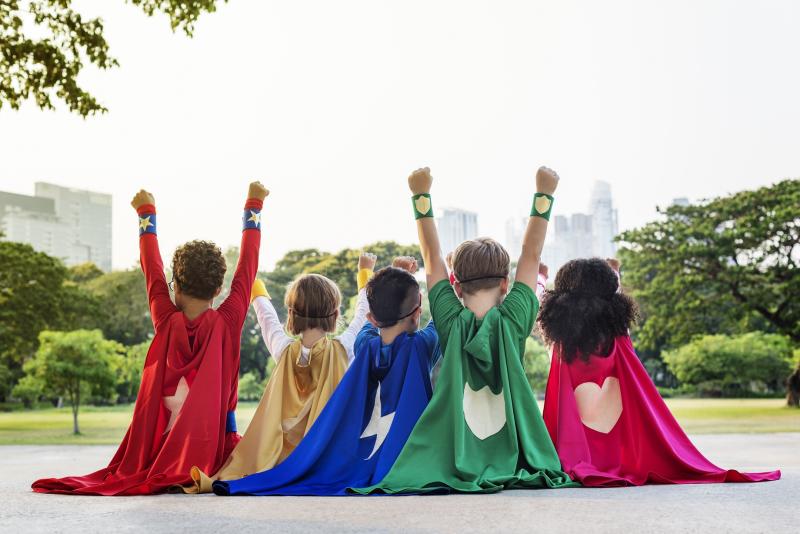 Five children dressed in superhero capes with their hands in the air as they look out to parkland