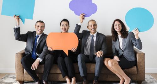 Four people sitting on a couch dressed in corporate attire holding up large speech bubble signs