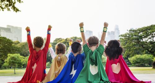 Five children dressed in superhero capes with their hands in the air as they look out to parkland