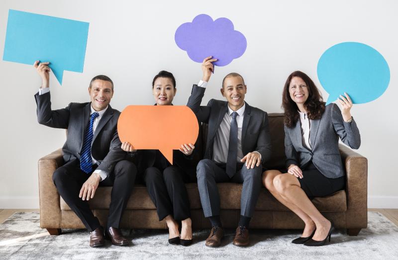 Four people sitting on a couch dressed in corporate attire holding up large speech bubble signs